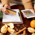 Młoda klientka trzyma kartę kredytową i smartfon nad stołem, zamierzając zamówić prezenty świąteczne dla rodziny