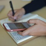 Przycięte ręce kobiety piszącej w notesie i używającej telefonu siedząc przy stole