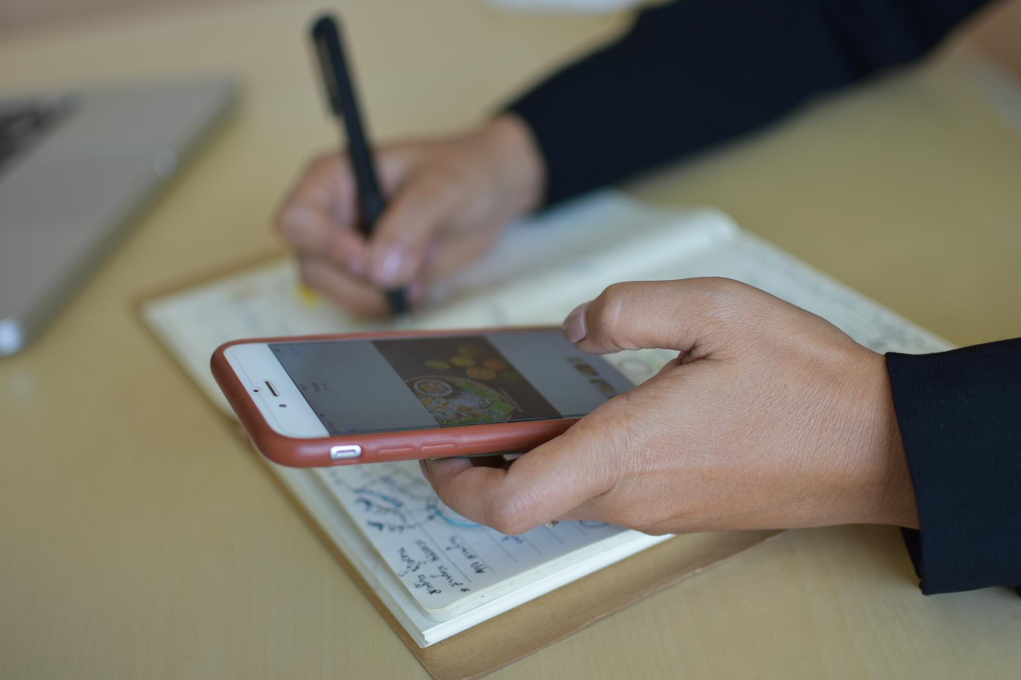 Przycięte ręce kobiety piszącej w notesie i używającej telefonu siedząc przy stole