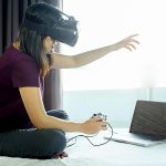 Kobieta z symulatorem wirtualnej rzeczywistości grająca w grę wideo na laptopie, siedząc w łóżku w domu