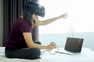 Kobieta z symulatorem wirtualnej rzeczywistości grająca w grę wideo na laptopie, siedząc w łóżku w domu