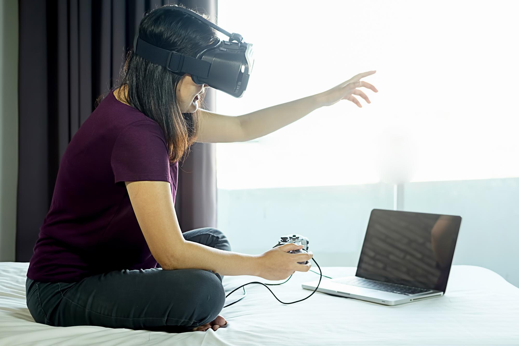 Kobieta z symulatorem wirtualnej rzeczywistości grająca w grę wideo na laptopie, siedząc w łóżku w domu