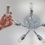 Ręka trzyma domową lampę kukurydzianą LED przed zamontowaniem w chromowanym żyrandolu sufitowym.