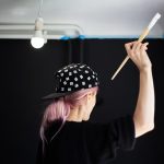 Widok młodej dziewczyny z różowymi włosami, malowanie ścian pędzlem i białym kolorem z tyłu, odświeżenie mieszkania. Nosi czarną czapkę i sweter.