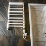 Biała nowoczesna elektryczna suszarka na ręczniki zamontowana na czarnej, wyłożonej kafelkami ścianie w nowoczesnej łazience.