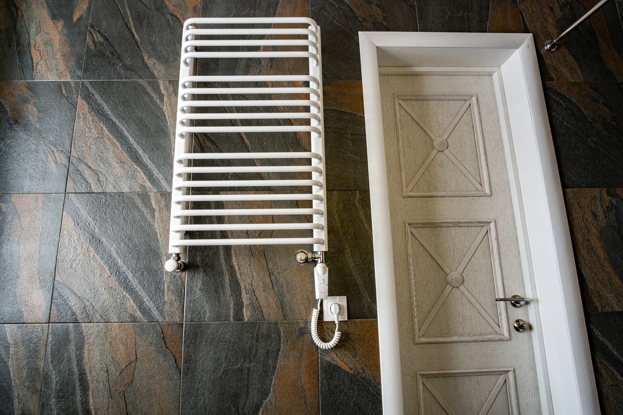 Biała nowoczesna elektryczna suszarka na ręczniki zamontowana na czarnej, wyłożonej kafelkami ścianie w nowoczesnej łazience.