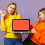 Dwie młode dziewczyny siostry bliźniaczki blondynka pokazując OK gest, przytrzymaj komputer PC z pustym pustym ekranem na białym tle na pastelowej fioletowej niebieskiej ścianie. Koncepcja życia rodzinnego osób. .