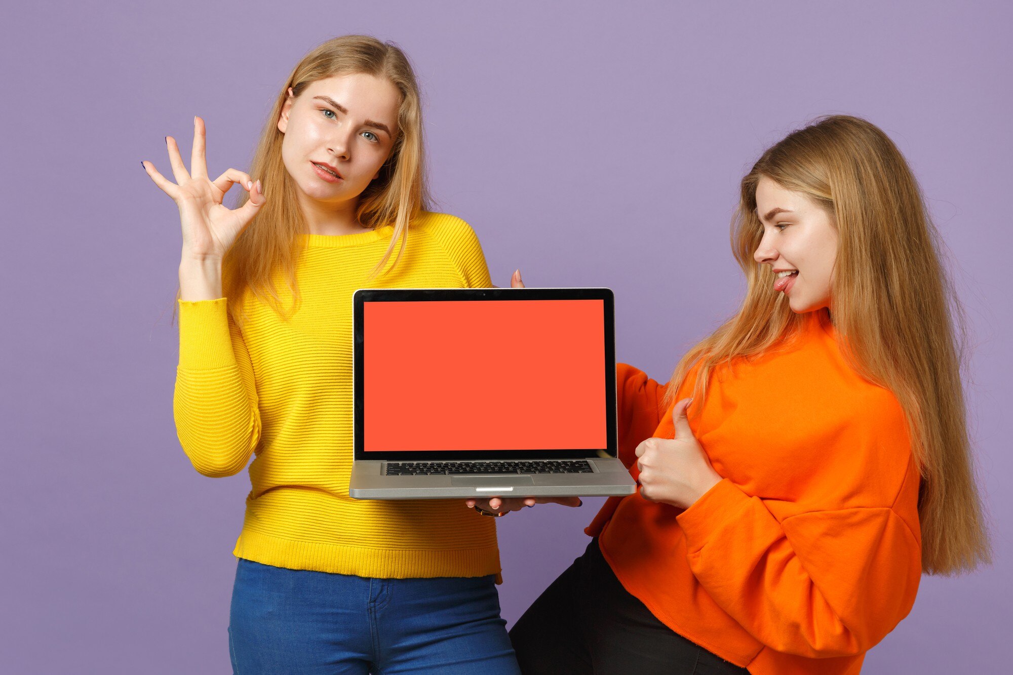 Dwie młode dziewczyny siostry bliźniaczki blondynka pokazując OK gest, przytrzymaj komputer PC z pustym pustym ekranem na białym tle na pastelowej fioletowej niebieskiej ścianie. Koncepcja życia rodzinnego osób. .