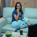 Kobieta trzyma pilota i je popcorn relaks na kanapie w salonie. Samotna szczęśliwa późno w nocy pani w piżamie odpoczywa przed telewizorem, leżąc na wygodnej kanapie, zmieniając kanały telewizyjne.