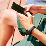 Ręce młodego sportowca w krótkich spodenkach przewijanie w telefonie komórkowym podczas relaksu w przerwie między treningami sportowymi na świeżym powietrzu