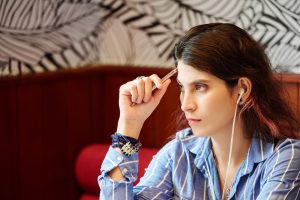 Zamyślona młoda kobieta słucha muzyki w słuchawkach i zastanawia się nad pomysłami na nowy projekt, siedząc przy kawiarnianym stoliku