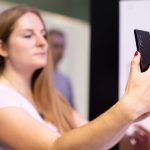 Młoda blondie dama trzyma w ręku czarny smartfon, zaglądając do niego, robiąc selfie c