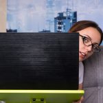 Młoda kobieta w okularach zerkająca zza laptopa, pracująca na laptopie, prowadząca interesy z domu, trzymająca długopis, gdy czyta informacje na ekranie