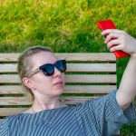 Blondynka robi sobie selfie, robi zdjęcie lub komunikuje się przez wideorozmowę przez telefon w słoneczny dzień w parku