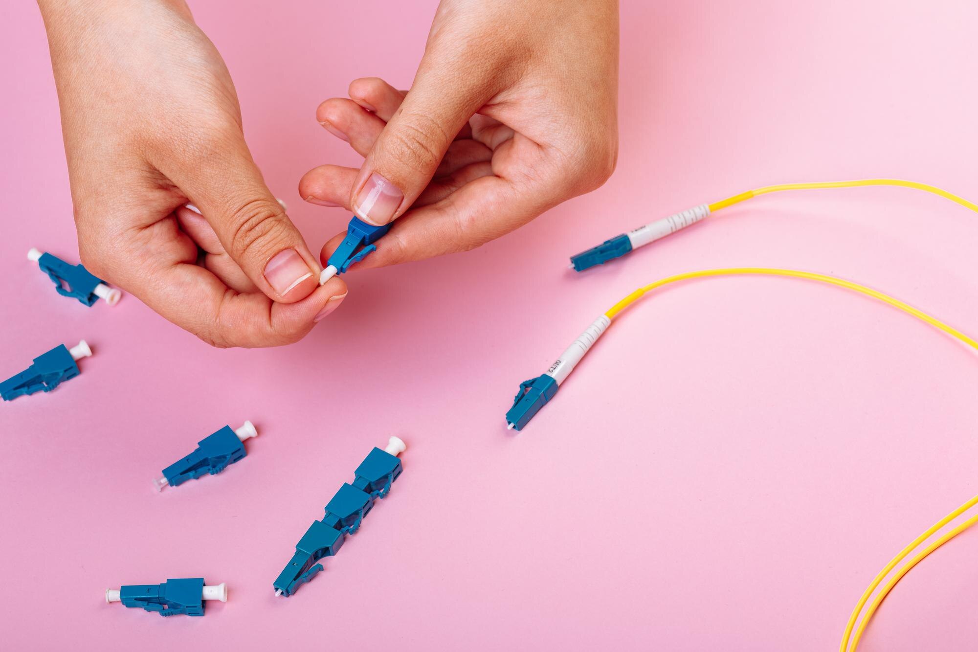 Ręka łączy niebieski tłumik optyczny LC na różowym tle.