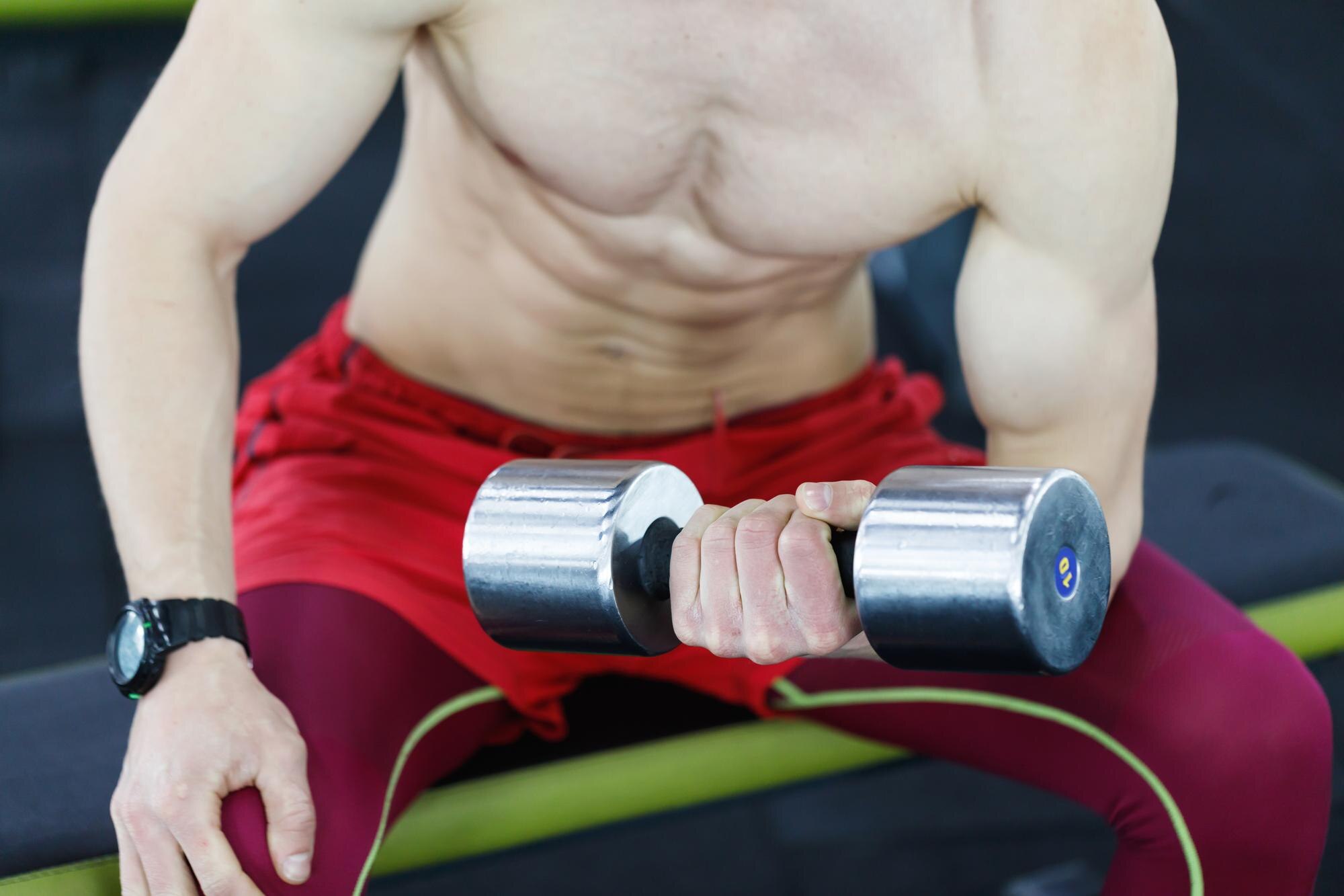 Zbliżenie na silnego mężczyznę topless siedzącego i podnoszącego hantle na siłowni