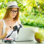 Fotografka wkłada lub wyjmuje kartę pamięci w swoim profesjonalnym aparacie dslr, gdy siedzi przy biurku z laptopem na tle rajskiej plaży zielonych palm