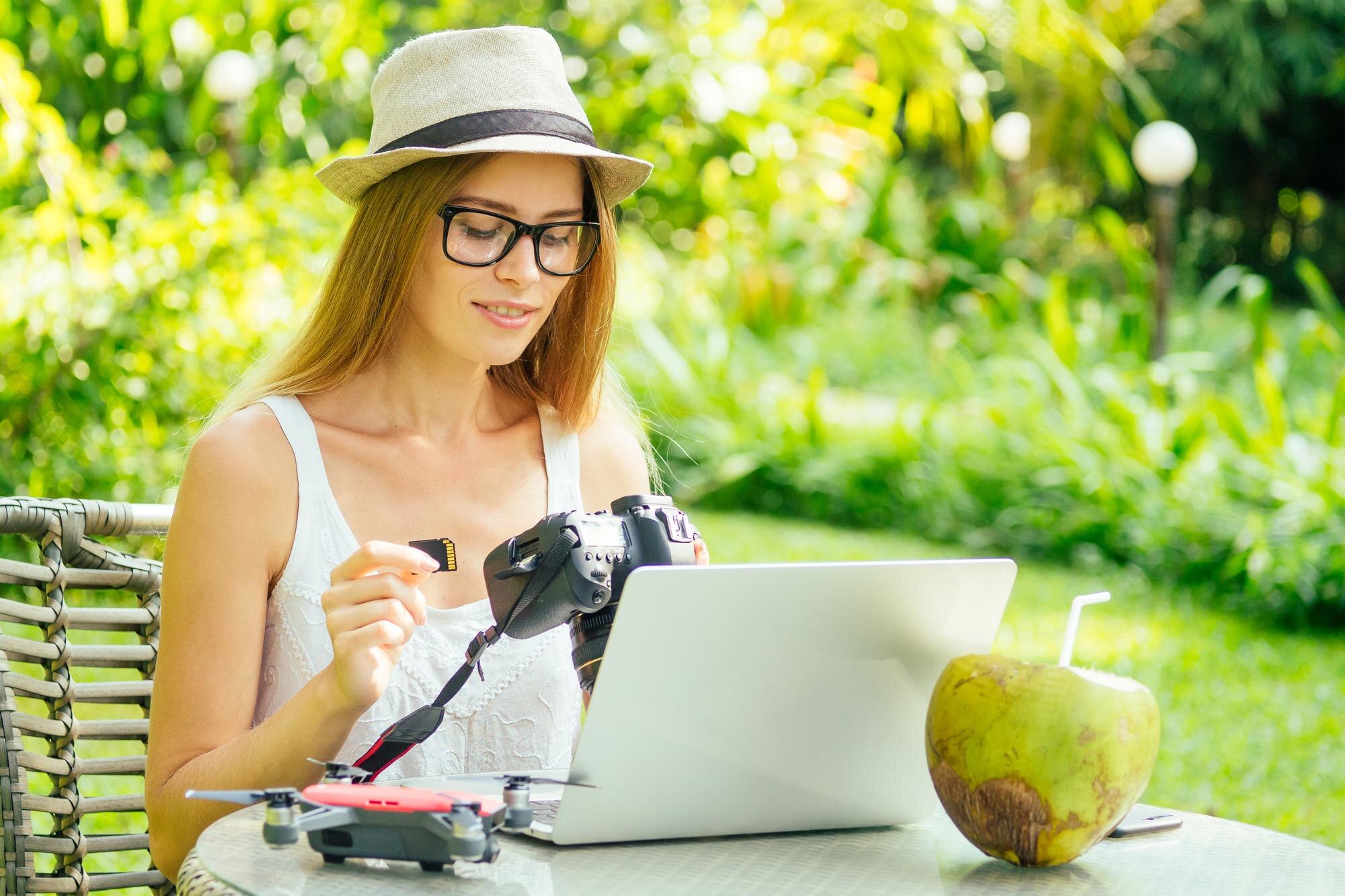 Fotografka wkłada lub wyjmuje kartę pamięci w swoim profesjonalnym aparacie dslr, gdy siedzi przy biurku z laptopem na tle rajskiej plaży zielonych palm