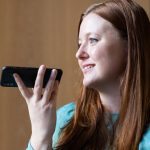 W biurze kobieta z smartfonem uśmiecha się i mówi do urządzenia, angażując się w rozmowę