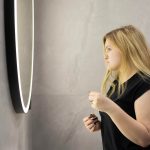 Młoda blondynka w czarnej koszuli badając jej odbicie w okrągłym oświetlonym lustrze w