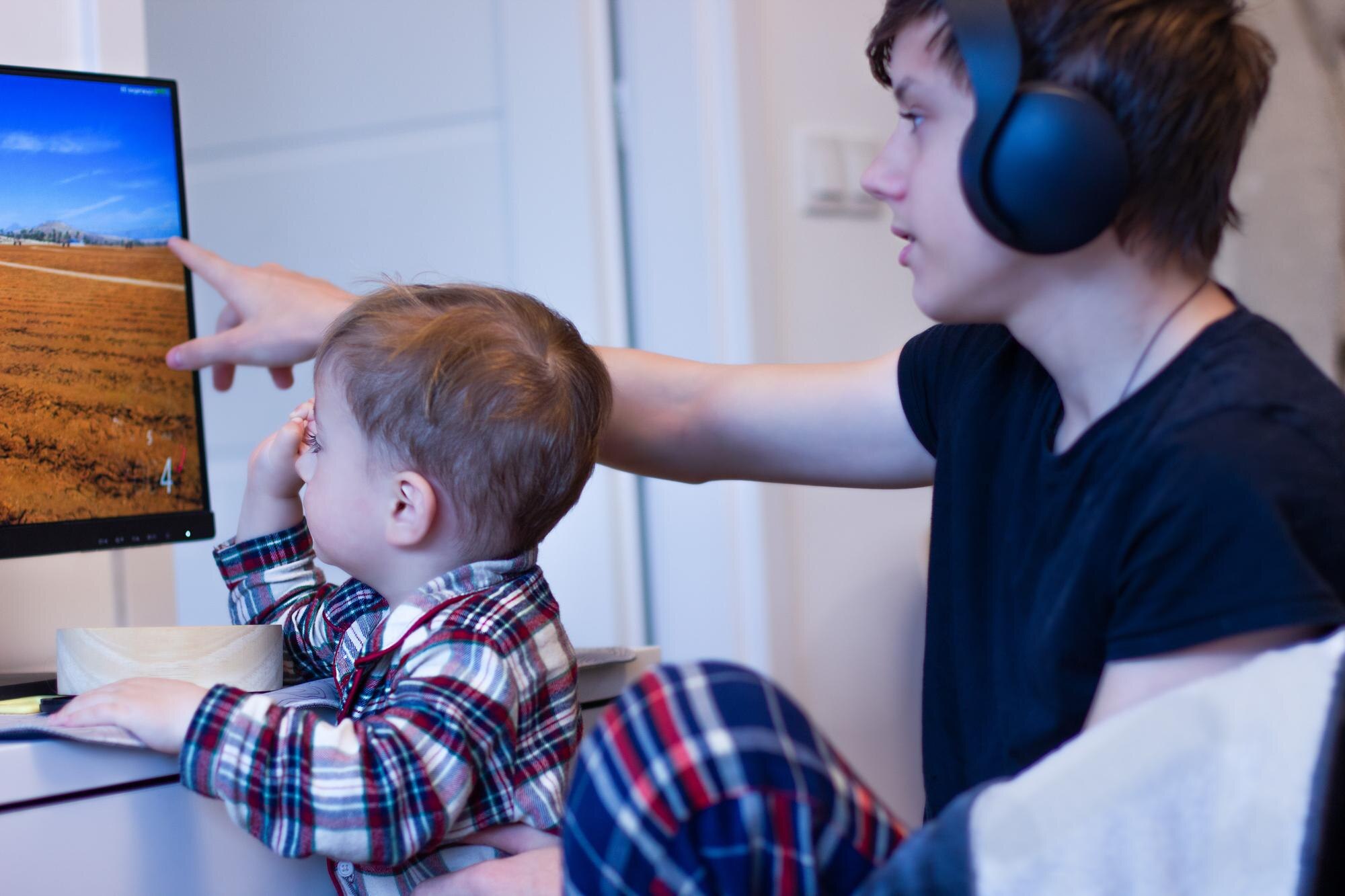 Chłopiec siedzi na ramionach nastolatka, a starszy brat pokazuje mu zdjęcie na monitorze.