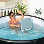 Dziewczyna w okularach przeciwsłonecznych radośnie macha ręką do ekranu laptopa w basenie na hotelowym tarasie