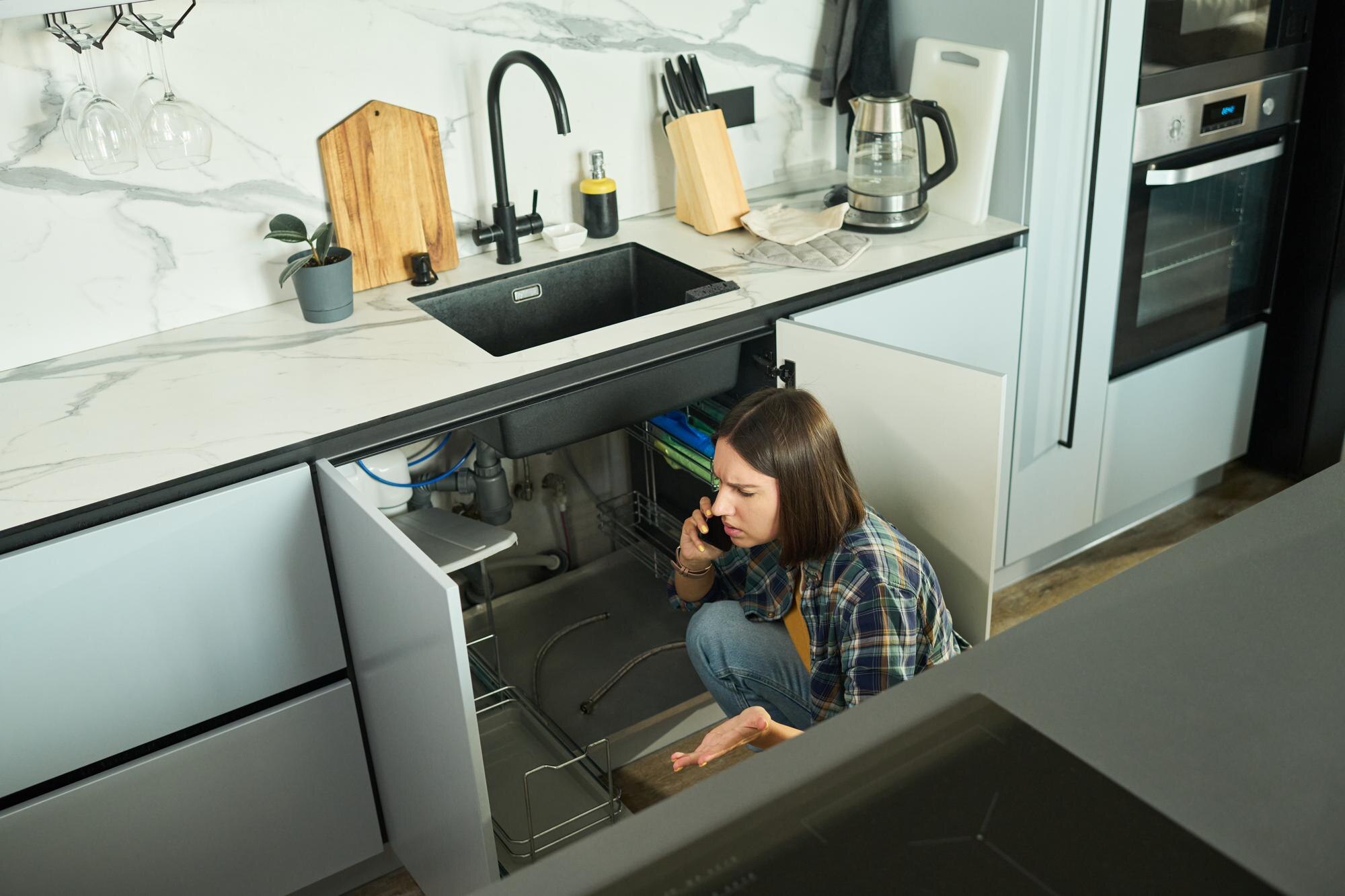Young Caucasian Woman Calling Plumber While Inspecting Leaking Kitchen Sink