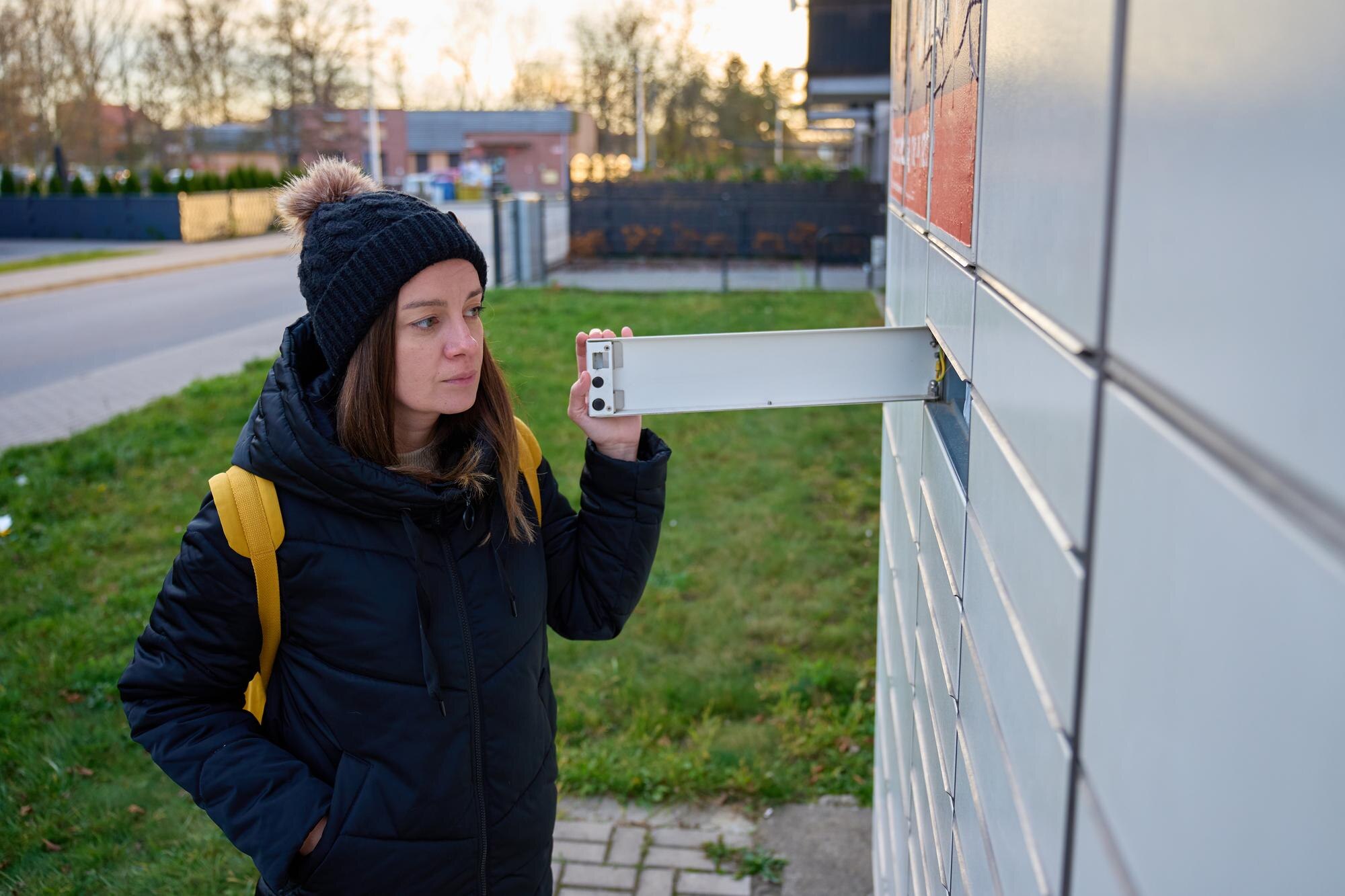 Woman wearing winter jacket and hat opening metal mailbox outside apartment building