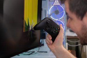 A person39s hand holds a black video game controller in front of a visibly cracked computer monitor representing gaming frustration and equipment damage