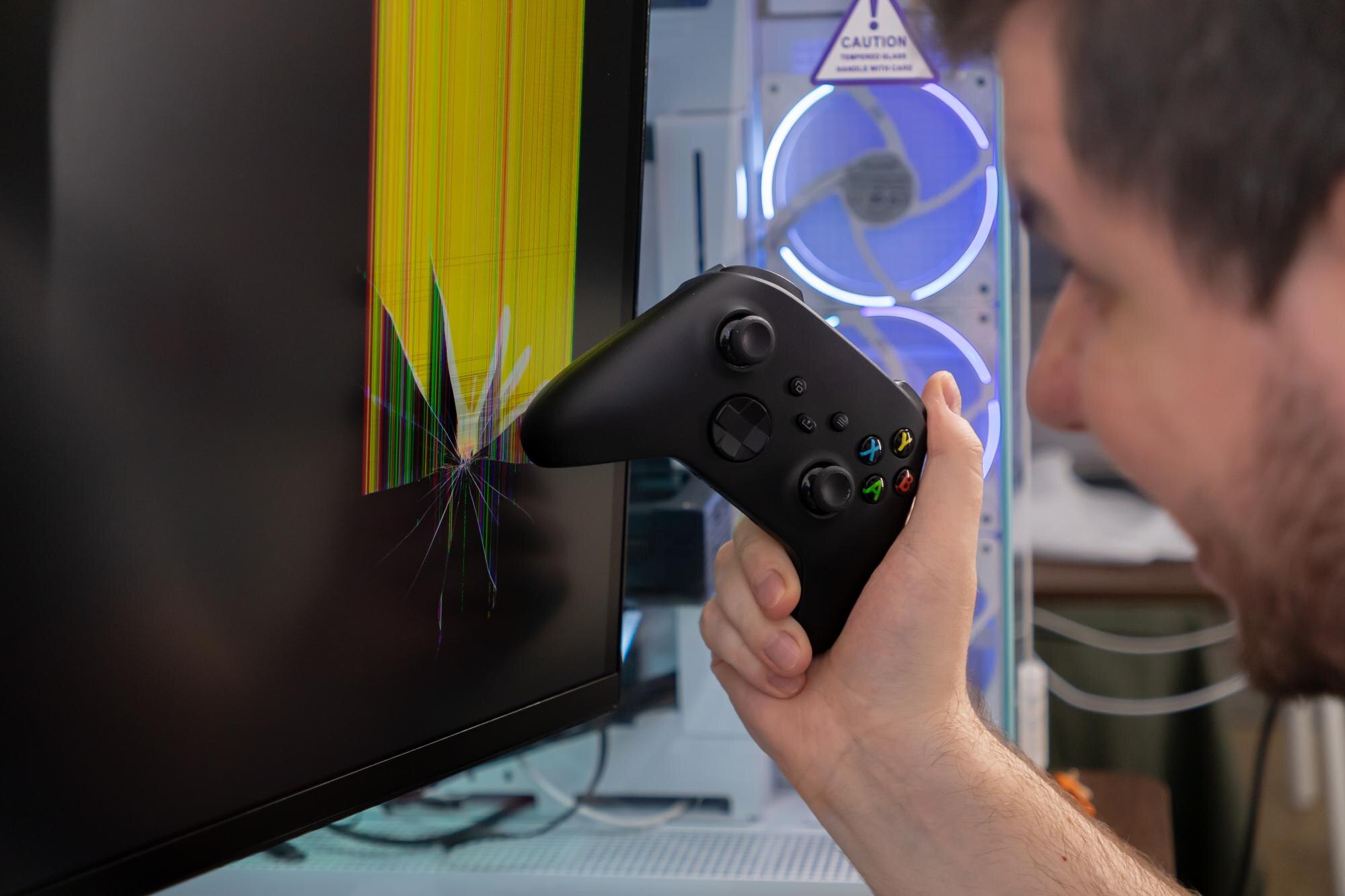A person39s hand holds a black video game controller in front of a visibly cracked computer monitor representing gaming frustration and equipment damage