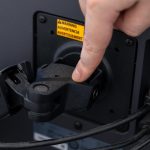 Hand adjusting a black monitor arm mount on a computer screen displaying a warning label illustrating ergonomic workstation setup and technology connectivity