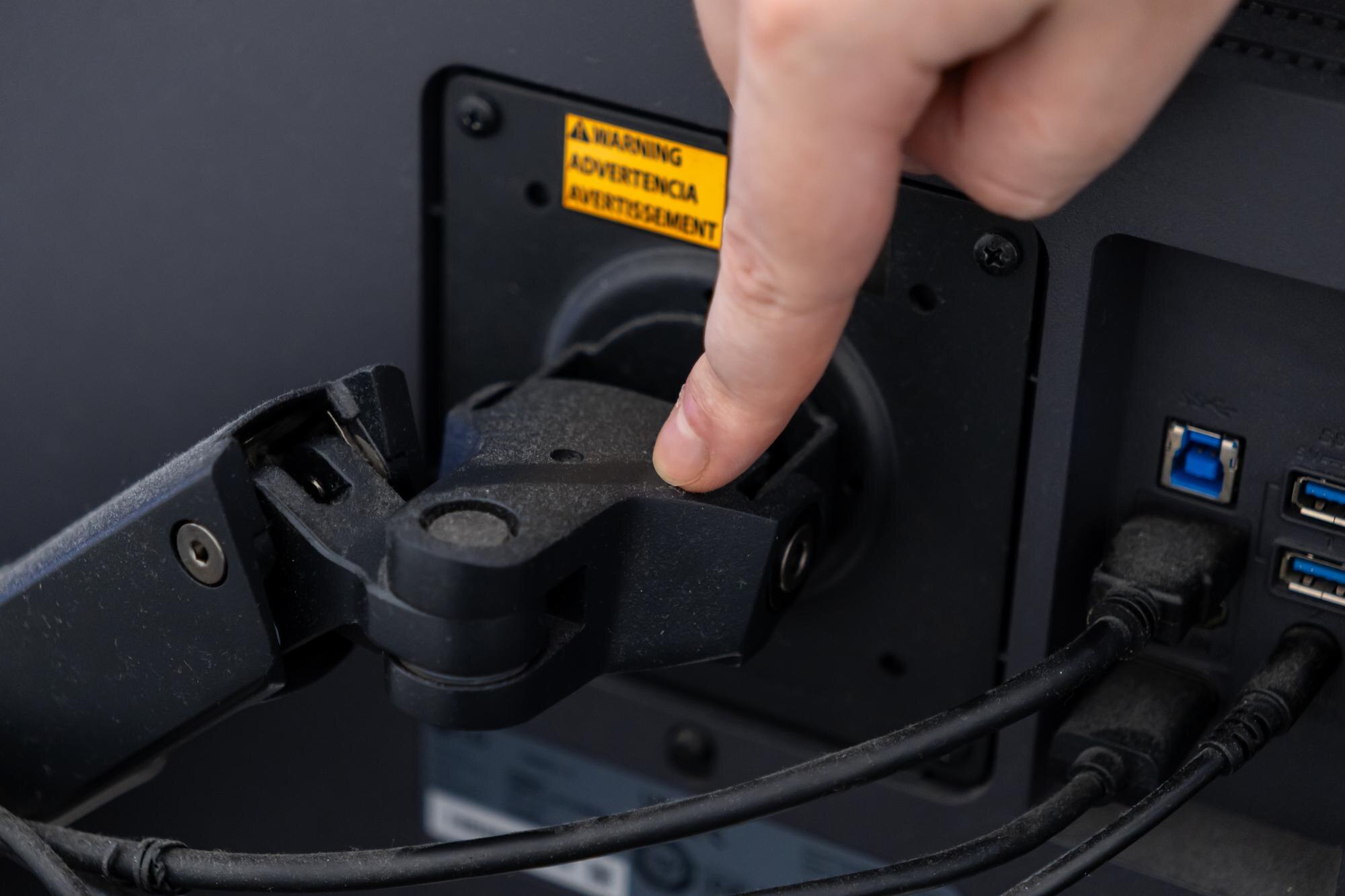 Hand adjusting a black monitor arm mount on a computer screen displaying a warning label illustrating ergonomic workstation setup and technology connectivity