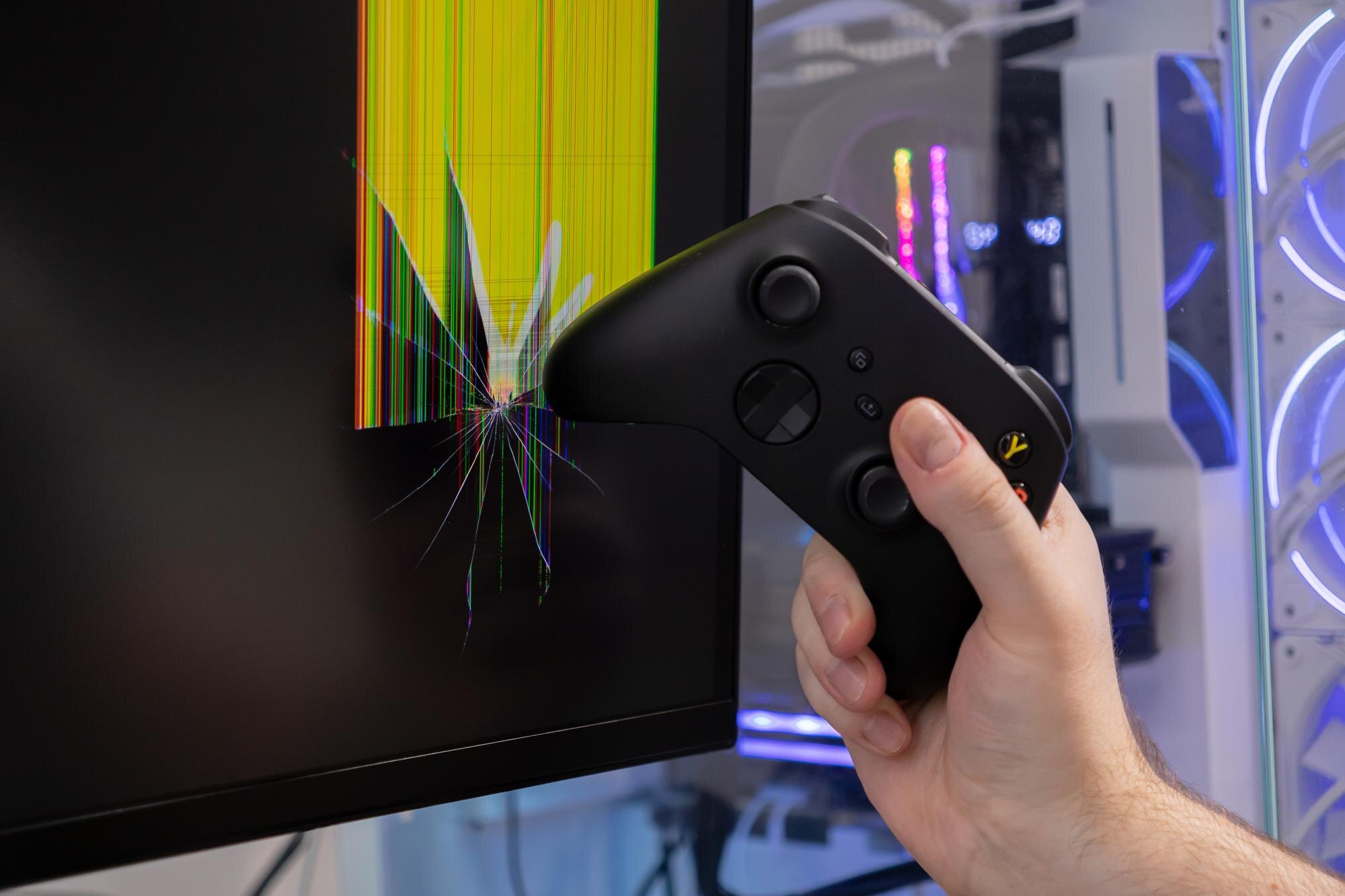 A gamer39s hand holds a black controller near a severely broken computer monitor depicting intense gaming frustration and damaged technology