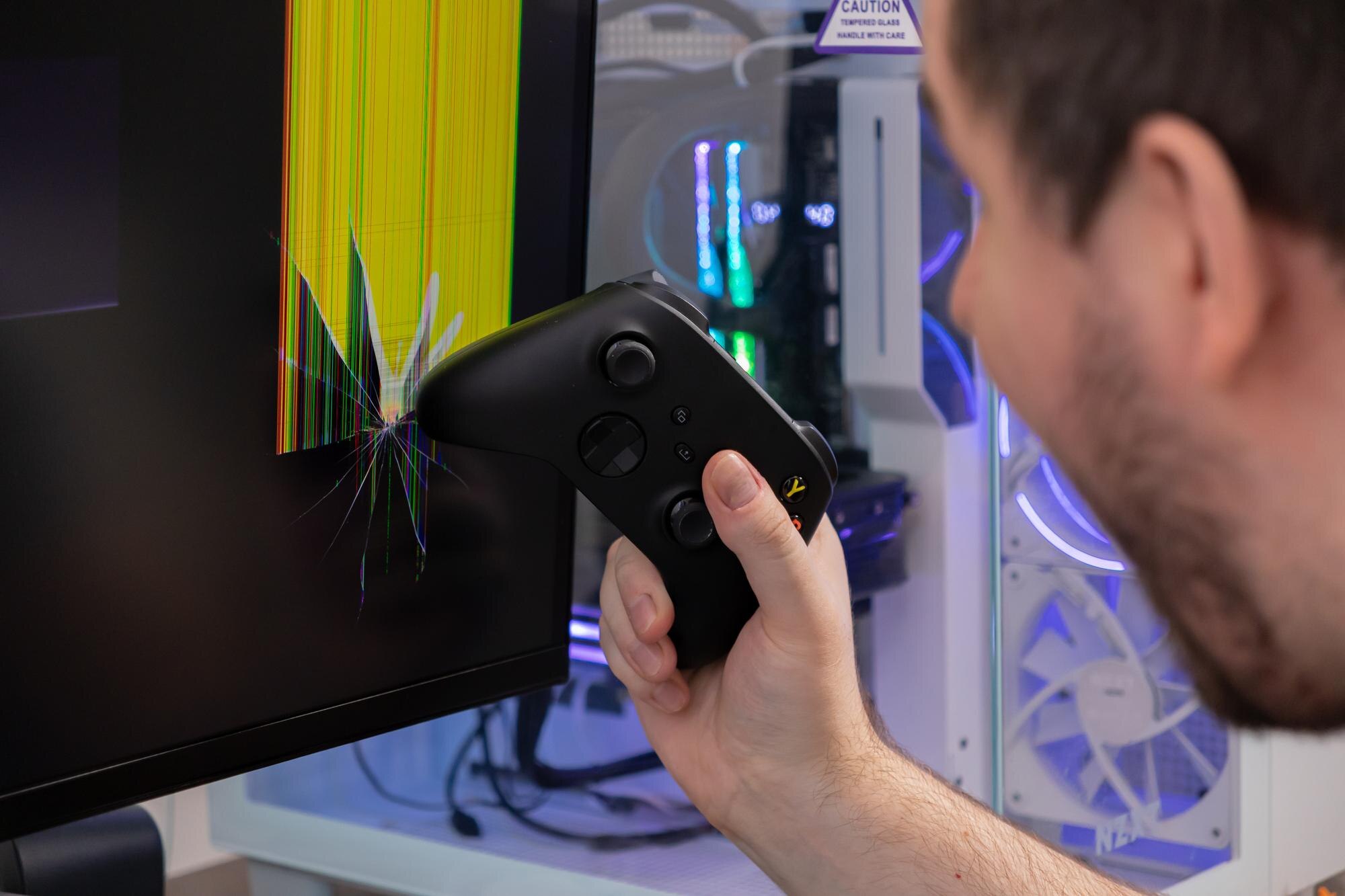Angry gamer holds black controller near severely cracked monitor displaying corrupted lines depicting gaming rage stress and technology damage