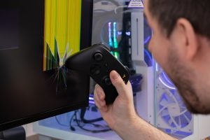 Angry gamer holds black controller near severely cracked monitor displaying corrupted lines depicting gaming rage stress and technology damage