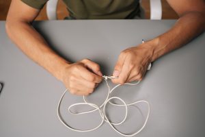 Closeup of technician hands tying knot in damaged white charger cable attempting repair at gray desk