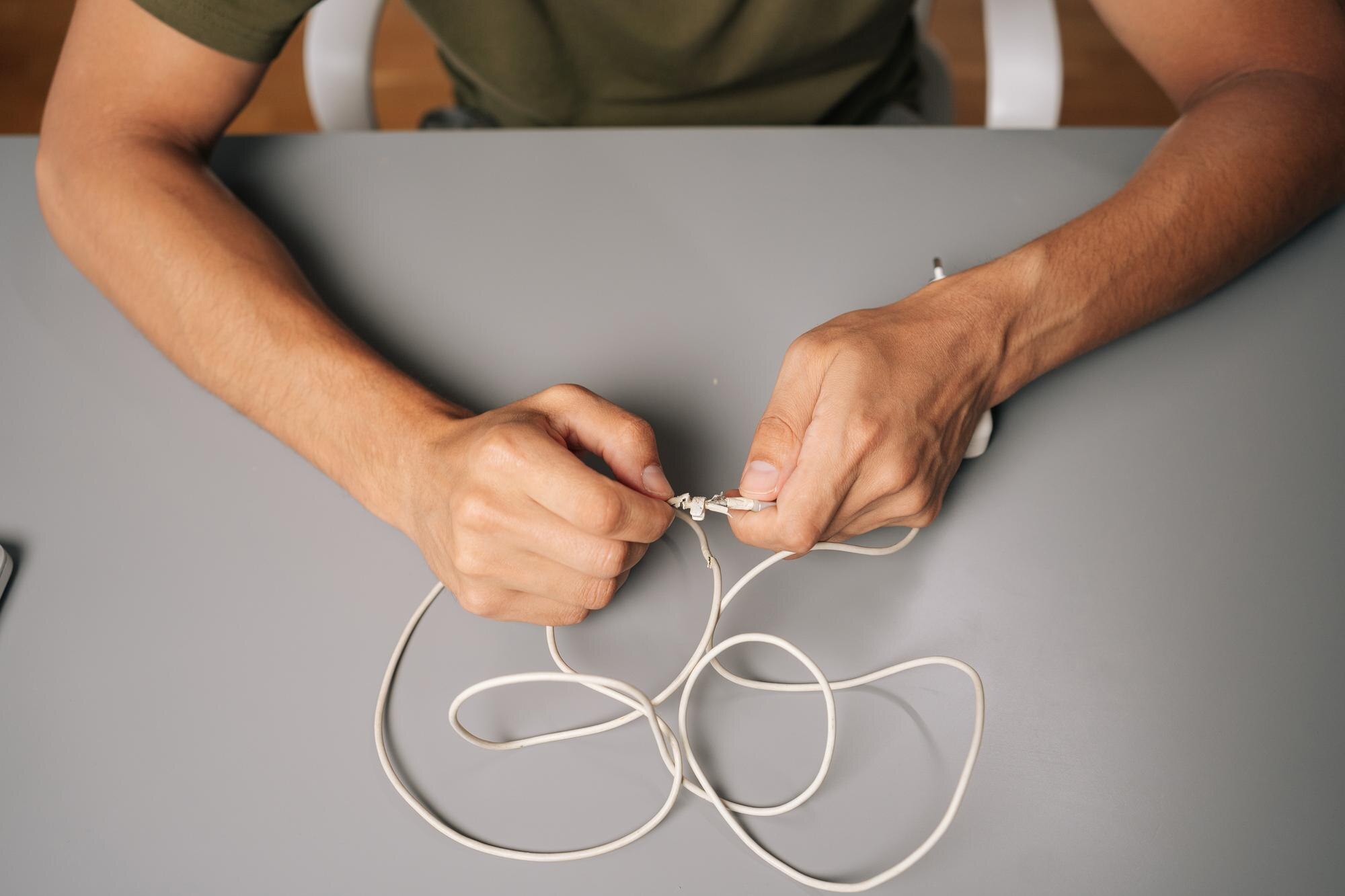 Closeup of technician hands tying knot in damaged white charger cable attempting repair at gray desk