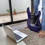 HOmbre, do którego widać tylko nogi, stojąc z gitarą elektryczną przed komputerem