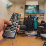 Osoba trzymająca pilota w pokoju z telewizorem w tle.