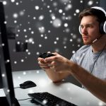 technologia, gry, rozrywka, zagrajmy i koncepcja ludzi - młody człowiek w zestawie słuchawkowym z gamepadem kontrolera, grając w gry komputerowe w domu i przesyłając strumieniowo grę lub wideo instruktażowe na śniegu