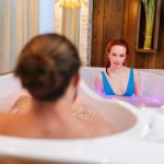 Widok z tyłu nie do poznania mężczyzny rozmawiającego z rudowłosą młodą kobietą w strojach kąpielowych siedzącą w podwójnej wannie z hydromasażem