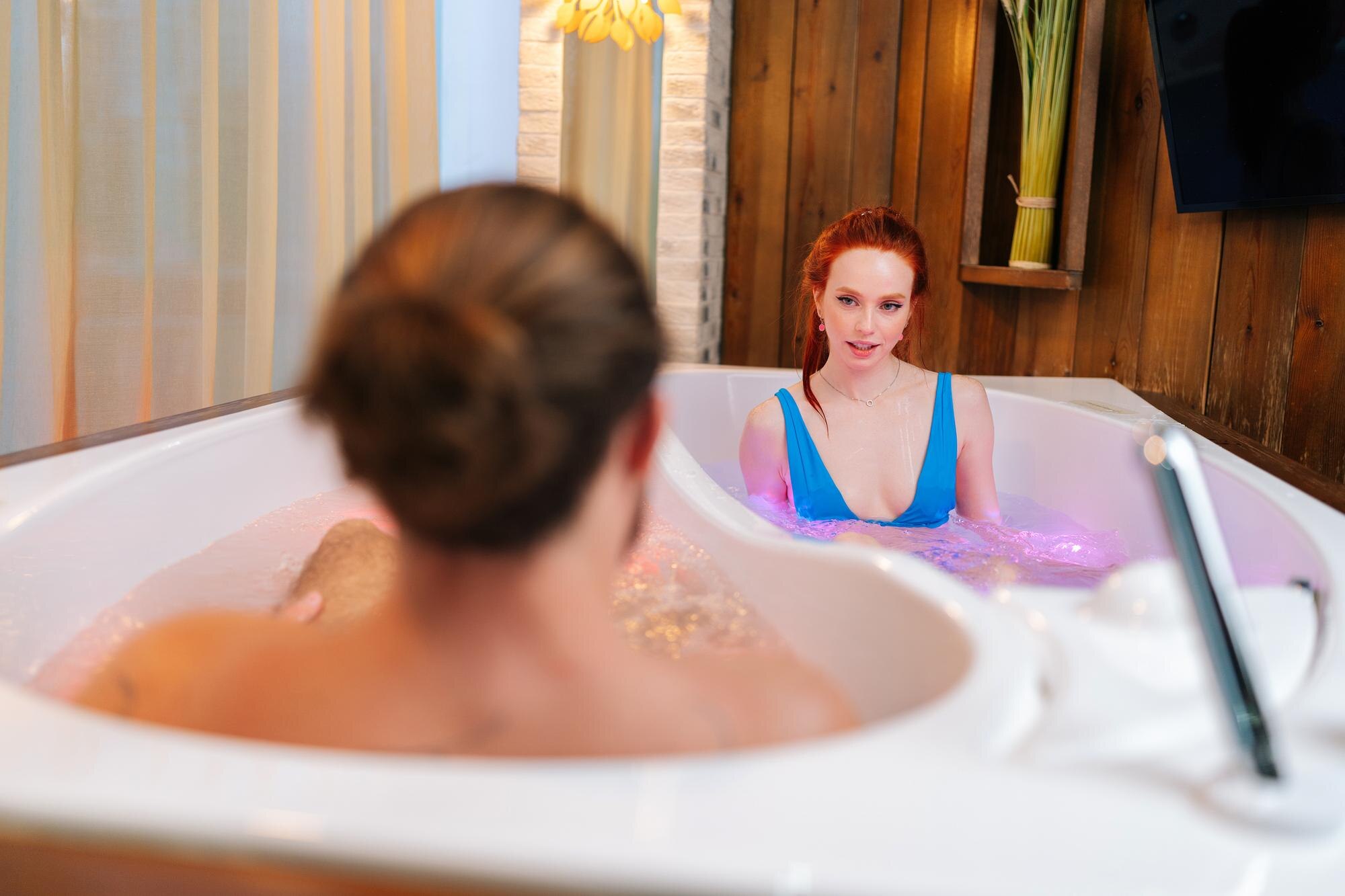 Widok z tyłu nie do poznania mężczyzny rozmawiającego z rudowłosą młodą kobietą w strojach kąpielowych siedzącą w podwójnej wannie z hydromasażem