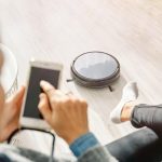 Przytnij nierozpoznawalną kobietę korzystającą ze smartfona i siedzącą na sofie w salonie z działającym odkurzaczem automatycznym