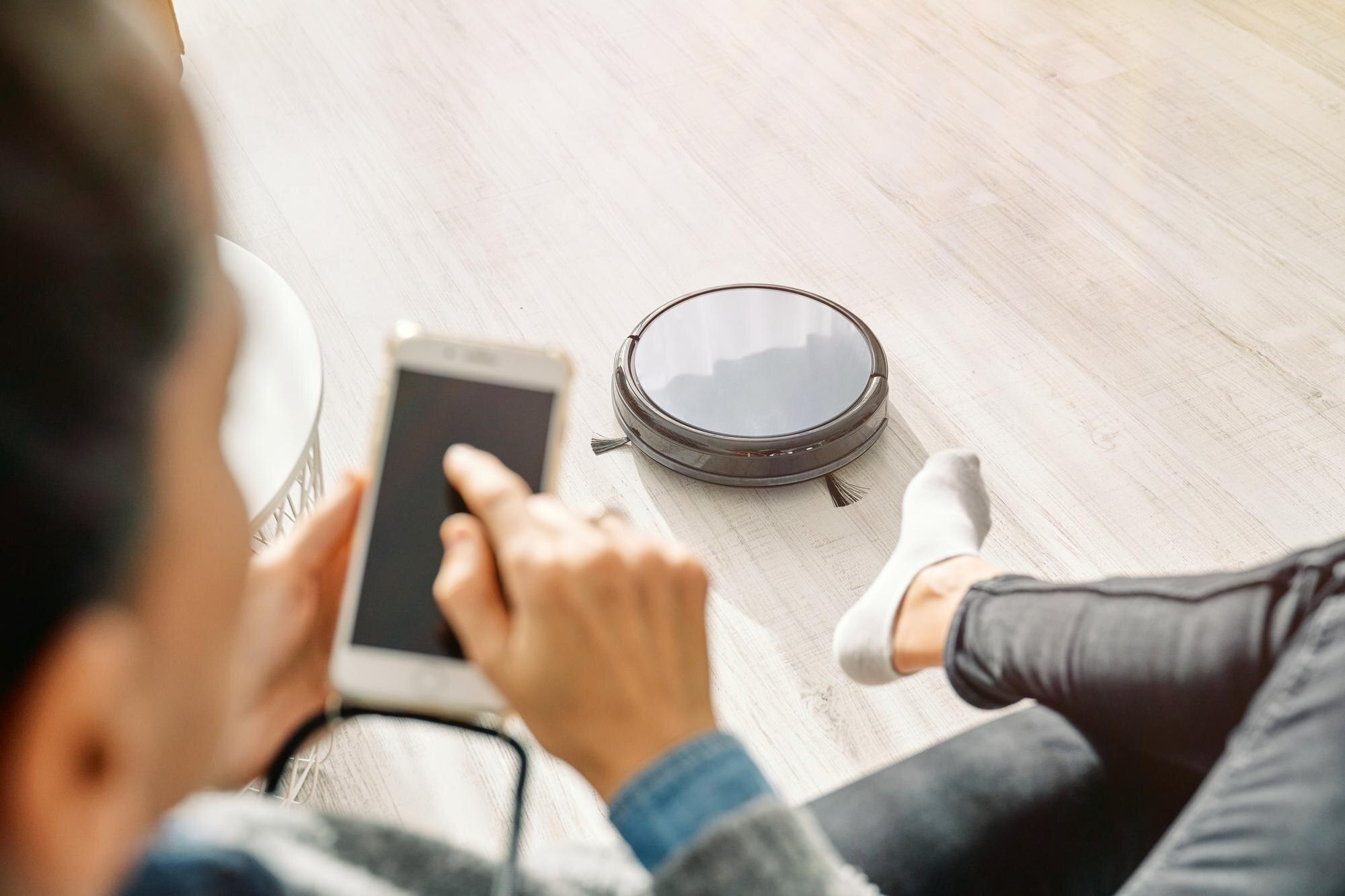 Przytnij nierozpoznawalną kobietę korzystającą ze smartfona i siedzącą na sofie w salonie z działającym odkurzaczem automatycznym