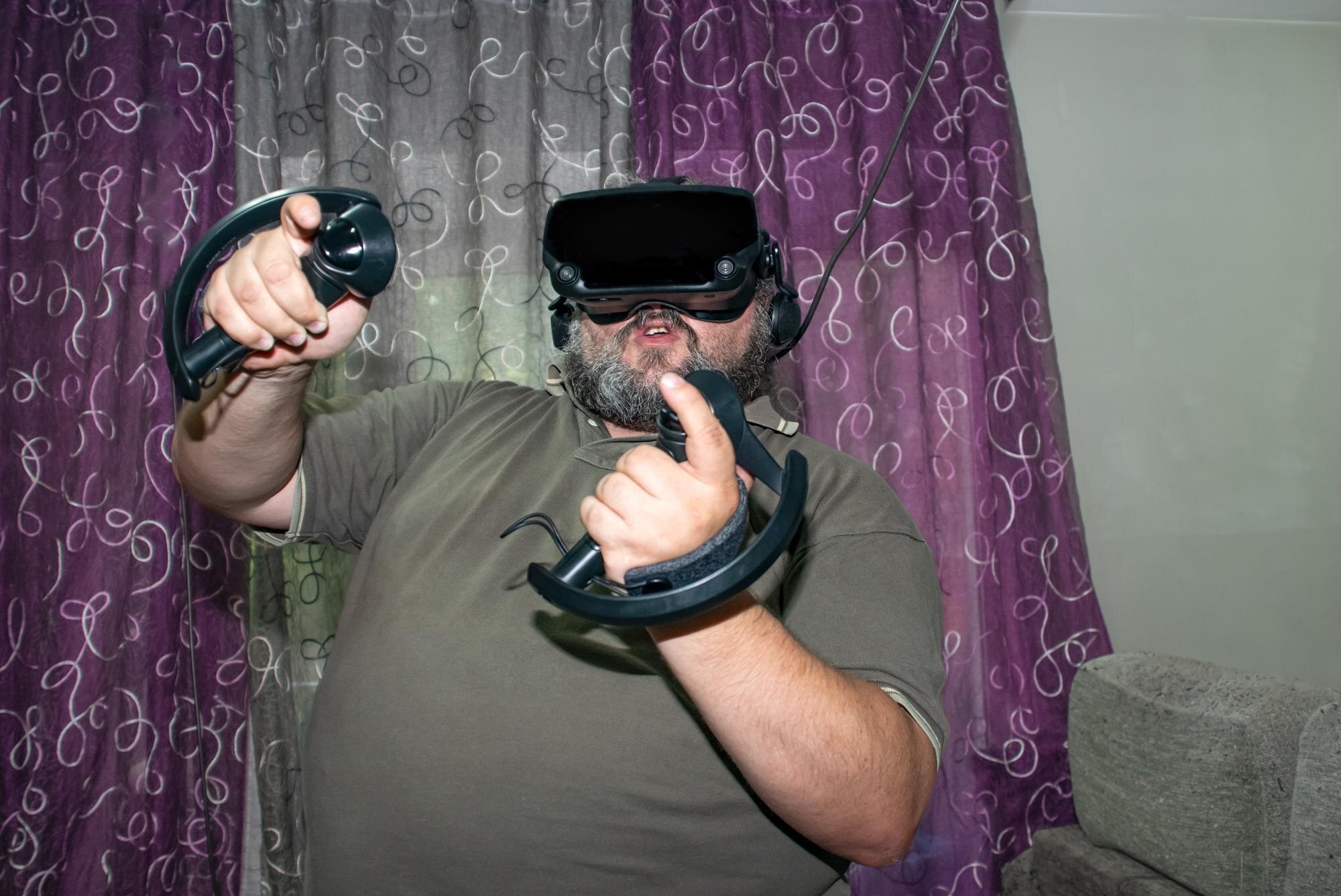 Nadwagi mężczyzna grający w wirtualną rzeczywistość podnoszący ręce z joystickami w VR z fioletowym i szarym tłem